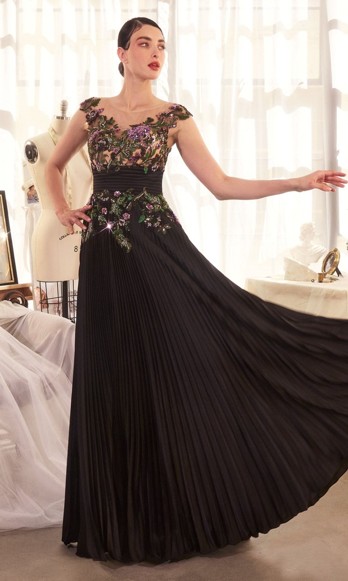 A woman poses indoors in the Andrea & Leo A1355 Sequin Long Pleated Prom Dress with floral appliques, standing near a dress form and sheer curtains in a bright room.